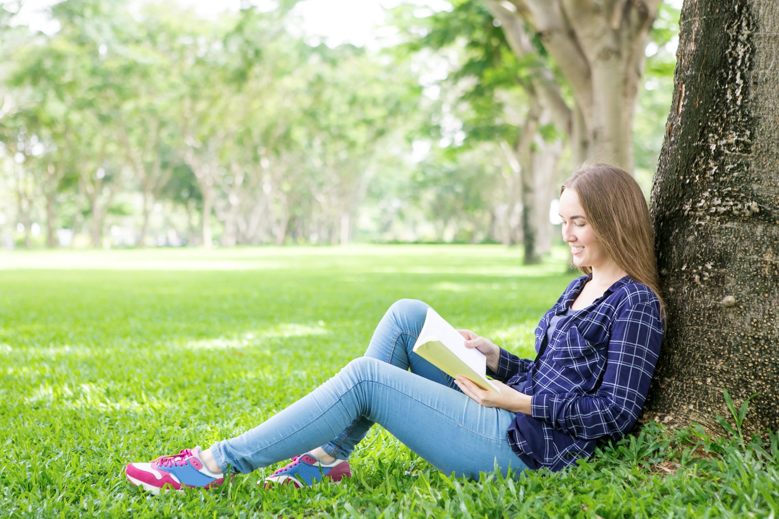 Positive student girl adoring reading book in summer park. Peaceful smiling young woman leaning on tree sitting on grass and having good time with favorite book. Learning outdoors concept